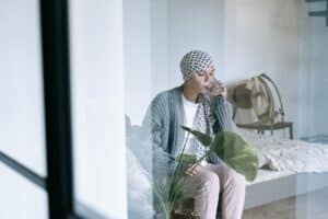 Woman in headscarf sitting on bed indoors, drinking water, significant in cancer awareness and treatment support.