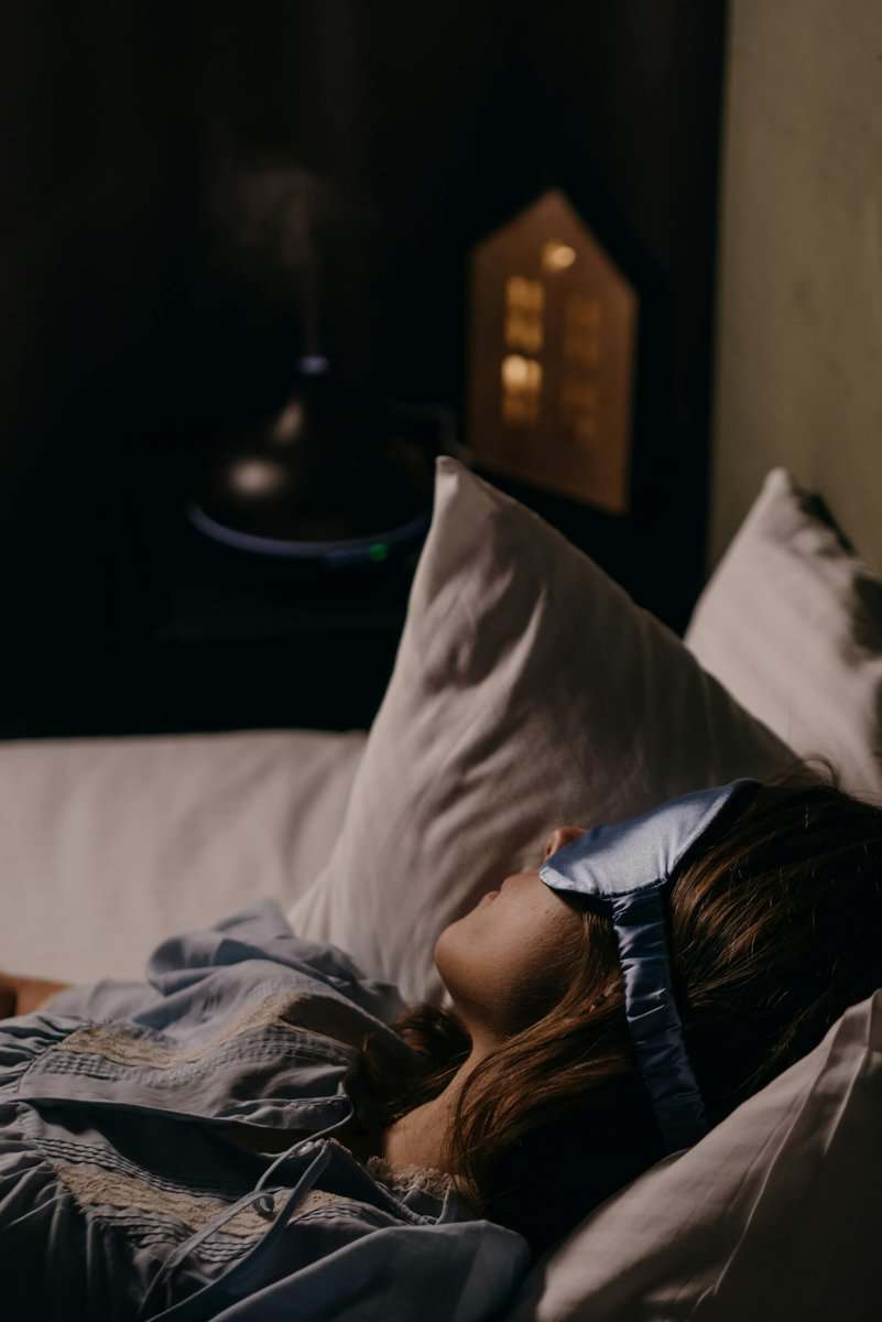A woman wearing a blue eye mask sleeping comfortably in a cozy bedroom setting.
