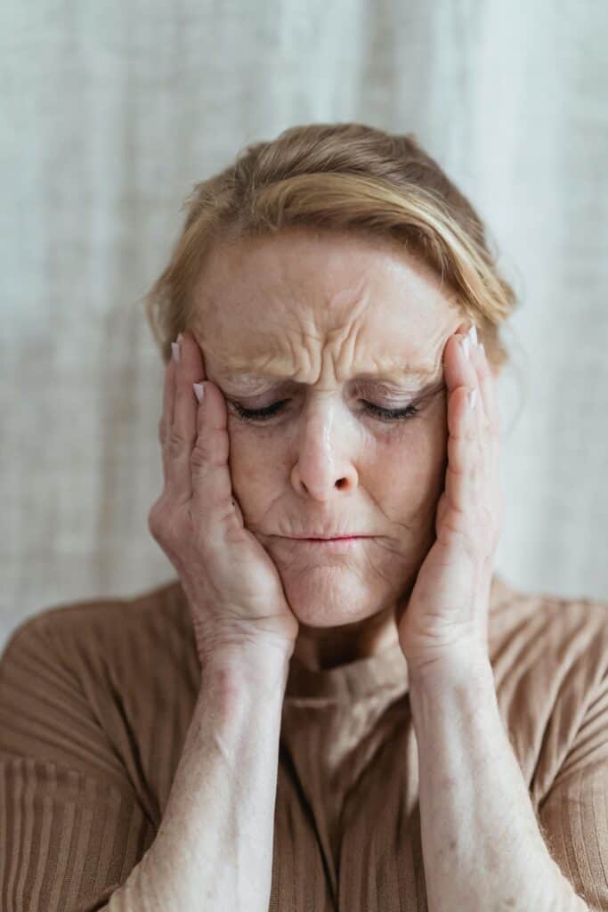 A senior woman with eyes closed, touching her face, showing signs of stress and concern indoors.