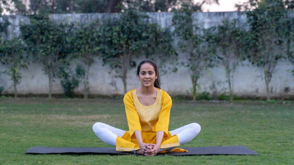 A young woman in a yellow shirt practices yoga in a garden, demonstrating a seated meditation pose.
